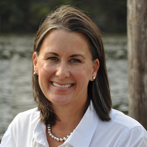 a head and shoulders portrait of an adult smiling with shoulder length brown hair wearing a white shirt and pearl necklace